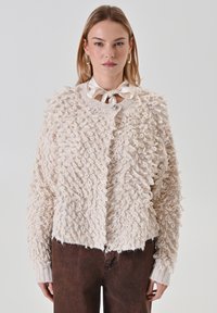 Cardigan crème texturé avec un motif en fil bouclé, col rond et fermeture à boutons. Stylisé avec un nœud de cou en satin clair. Pantalon marron visible.