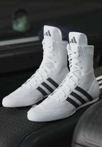 White high-top athletic shoes with black three-stripe branding, textured fabric, flat sole, and lace-up front detail.