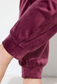 Burgundské sametové tepláky s elastickými manžetami, které se pyšní hladkým povrchem a mírným nařasením u kotníku.