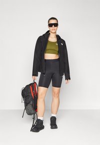 Fekete vízálló dzseki, olíva zöld sportmelltartó, fekete kerékpáros short. Fekete és piros hátizsák az egyik vállon, vastag fekete cipők, oversized napszemüveg.