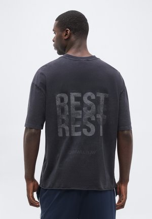 Bărbat cu fața spre spate, purtând un tricou negru cu text dublu estompat „BEST REST” pe spate, pe un fundal simplu și deschis la culoare.