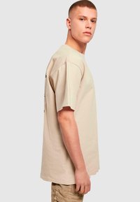 Beige bomull t-skjorte med korte ermer, avslappet passform, subtilt merkevarelogo på baksiden, vist på en modell som er vendt litt til siden.