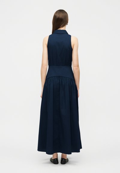 Γυναίκα με πλάτη, φορώντας αμάνικο μακρύ φόρεμα σε σκούρο μπλε με εφαρμοστή μέση και μαύρα επίπεδα παπούτσια, μπροστά σε λευκό, απλό φόντο.