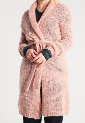 Frau trägt eine weiche, rosa Strickjacke mit Gürtel in der Taille, darüber ein dunkles Langarmshirt und dunkle Hosen.