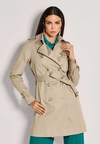 Vrouw met lang donker haar, gekleed in een beige double-breasted trenchcoat, die om de taille is gebonden over een teal shirt en broek, poseert met haar hand op de kraag.