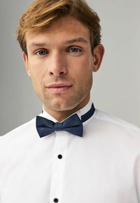 Weißes Hemd mit schwarzen Knöpfen, ausgestattet mit einer blauen Karottenfliege. Glatter Stoff mit strukturiertem Kragen, minimalistisches Design.