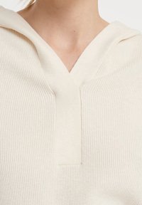 Maglione a coste color crema con scollo a V e cappuccio. La texture sembra morbida, presenta un design semplice e un dettaglio centrale con cucitura frontale.