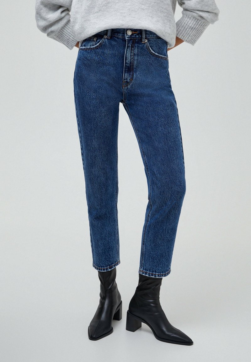 Personne portant un jean bleu taille haute, un pull gris et des bottines noires à talons, debout les mains sur les hanches devant un fond uni.