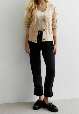 Kobieta ubrana w beżowy kardigan z grubym splotem na białym topie, czarne spodnie z mankietami, czarne mokasyny i warstwowe złote naszyjniki, stojąca na tle jednolitej ściany.