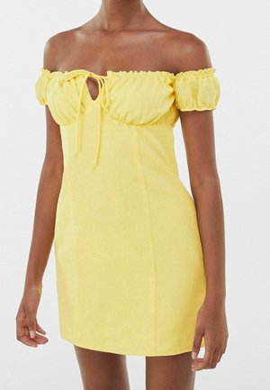 Vrouw draagt een korte, gele jurk met blote schouders, pofmouwen en een strikdetail aan de voorkant bij de borst.