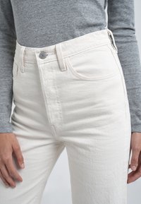 Personne portant un jean blanc taille haute avec un bouton et une fermeture éclair, associé à un haut gris à manches longues, mains reposant le long du corps.