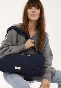 Sac à épaule en feutre bleu avec une poche zippée, doté d'une bandoulière bleu marine. Le modèle porte un pull en laine gris et un jean.