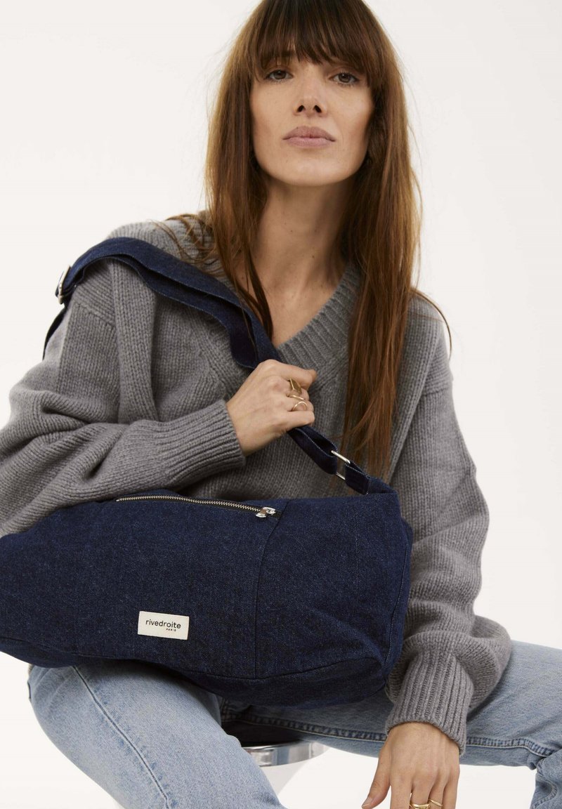 Sac à épaule en feutre bleu avec une poche zippée, doté d'une bandoulière bleu marine. Le modèle porte un pull en laine gris et un jean.