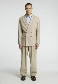 Bež saka s dvojim gumbom z teksturirano površino, ki ima velike temne gumbe. V kompletu s svetlo modro srajco s ovratnikom in rjavimi čevlji.