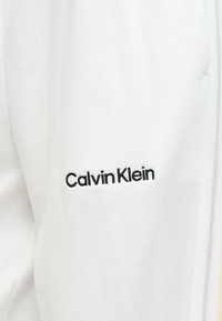 Λευκό ύφασμα με μαύρο κεντημένο λογότυπο "Calvin Klein" σε ρούχο, που δείχνει μέρος ραφής φερμουάρ στα δεξιά.