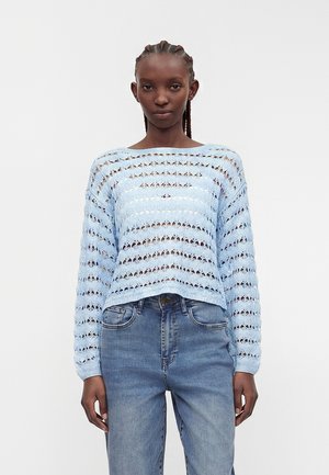 Kvinde med fletninger iført en lyseblå åbenstrikket sweater og blå højtaljede jeans, stående foran en ensfarvet hvid baggrund.