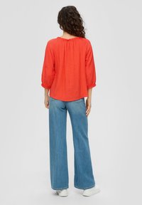 Blouse rouge texturée avec des manches bouffantes trois-quarts, associée à un jean large bleu clair et des baskets blanches.