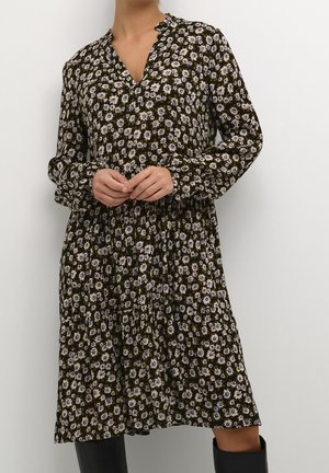 Woman wearing a long-sleeve, knee-length dress with a dark floral pattern and black knee-high boots against a plain background.