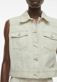 Chaleco de denim sin mangas de color beige claro con costuras verdes, cuatro bolsillos frontales y seis botones en el cierre. Textura suave.