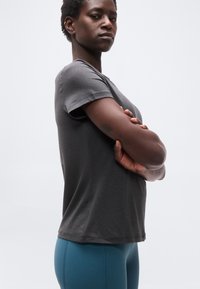 Αυτός/Αυτή που φαίνεται σίγουρος/η, με διασταυρωμένα χέρια, φοράει γκρι κοντομάνικο πουκάμισο και μπλε κολάν, στέκεται μπροστά σε απλό λευκό φόντο.