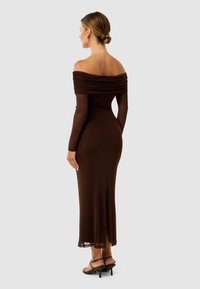 Robe marron dénudée sur les épaules avec des détails froncés, manches longues, silhouette ajustée et fente latérale, accompagnée de sandales à talons.