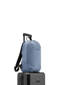 Un sac à dos bleu avec une texture lisse, doté de bretelles réglables et d'un design minimaliste, reposant sur une valise noire à rainures rigides.