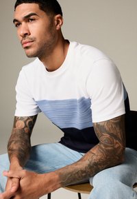 Kortärmad t-shirt i vitt, ljusblått och marinblå ränder. Bomullsmaterial med rund halsringning. Framträdande tatueringar på armarna. Jeans i ljus tvätt.