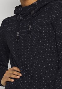 Sudadera azul marino con un patrón texturizado de puntos y costuras en zigzag. Cuenta con cordones ajustables y un bolsillo frontal.