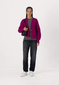 Cardigan a maglia fucsia con maniche a costine, t-shirt grigia, jeans dritti scuri e sneakers bianche; borsa a tracolla nera con logo.