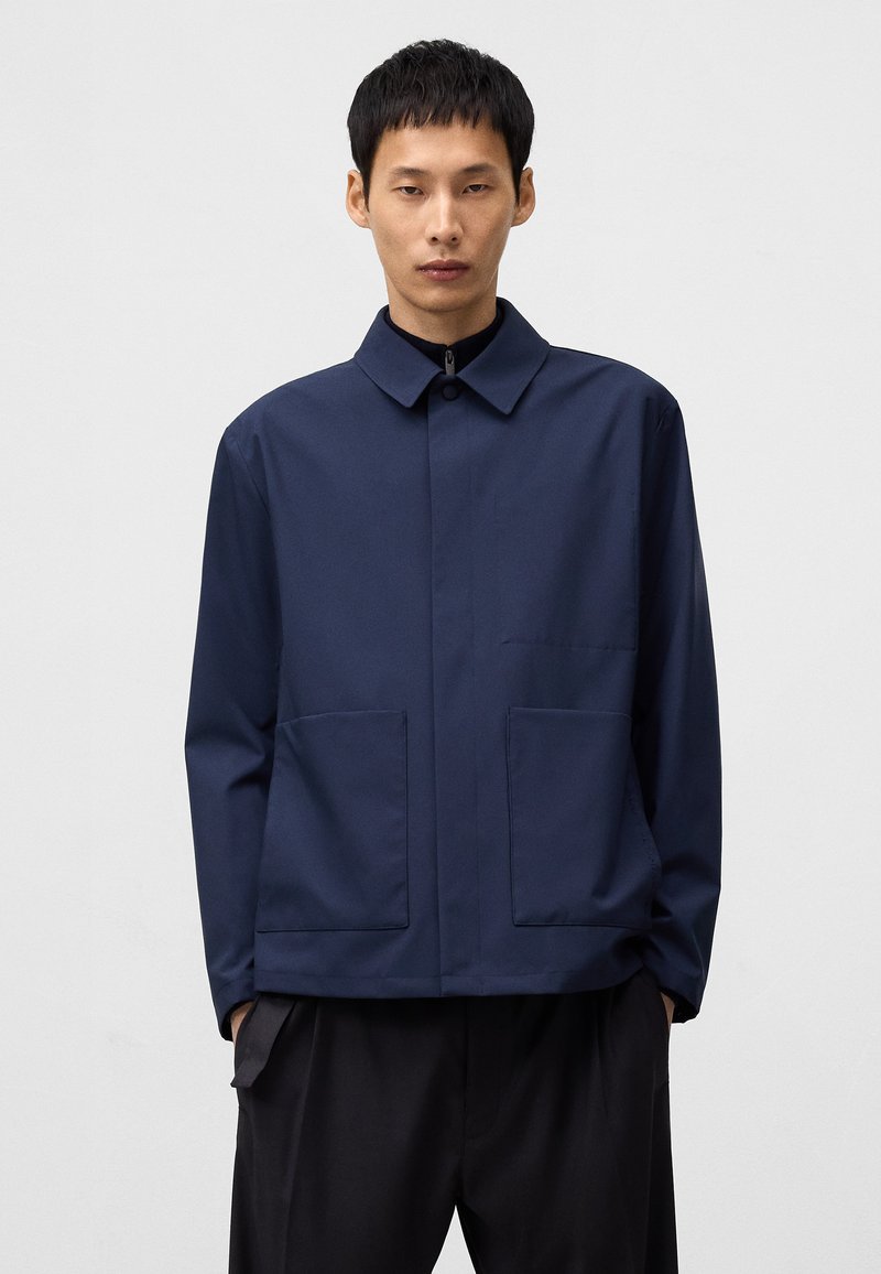 Granatowa kurtka z kołnierzykiem, z zapięciem na guziki z przodu, dwiema dużymi kieszeniami naszytymi oraz luźnym krojem. Gładki, teksturowany materiał.