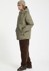 Doudoune verte olive avec capuche, dotée de poches latérales et d'une poche poitrine, associée à un pantalon en velours côtelé marron et des bottes noires à lacets.