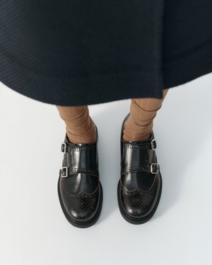 Czarne skórzane buty brogue z podwójnymi klamrami, noszone z brązowymi skarpetkami i ciemną spódnicą na białym tle.