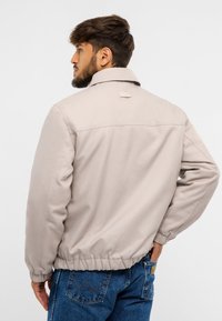 Beżowa kurtka z kołnierzykiem, elastycznym pasem i gładkim materiałem. Charakteryzuje się minimalistycznym wzornictwem i małą metką z tyłu.