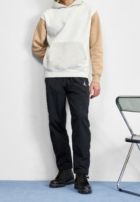 Felpa grigia chiara e marrone con tasca frontale, abbinata a pantaloni neri e stivali neri con lacci. Presenta un logo sui pantaloni.