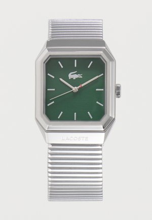 Ceas metalic argintiu cu cadran pătrat verde, marcatori de oră argintii, logo Lacoste și baretă metalică zimțată. Ora indicată este 10:11:38.