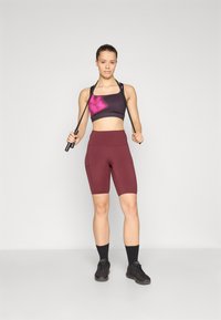 Sportski outfit s crno-ljubičastim tie-dye sportskim grudnjakom i crvenim visoko-strukiranim biciklističkim šortsevima, upotpunjen crnim sportskim cipelama i čarapama.