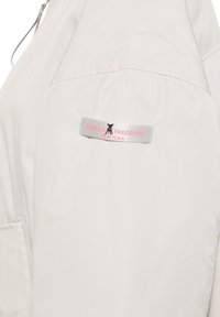 Lichtbeige jack gemaakt van een gladde stof, met een logo-opdruk in grijs en roze op de linkerschouder en een ritsluiting.