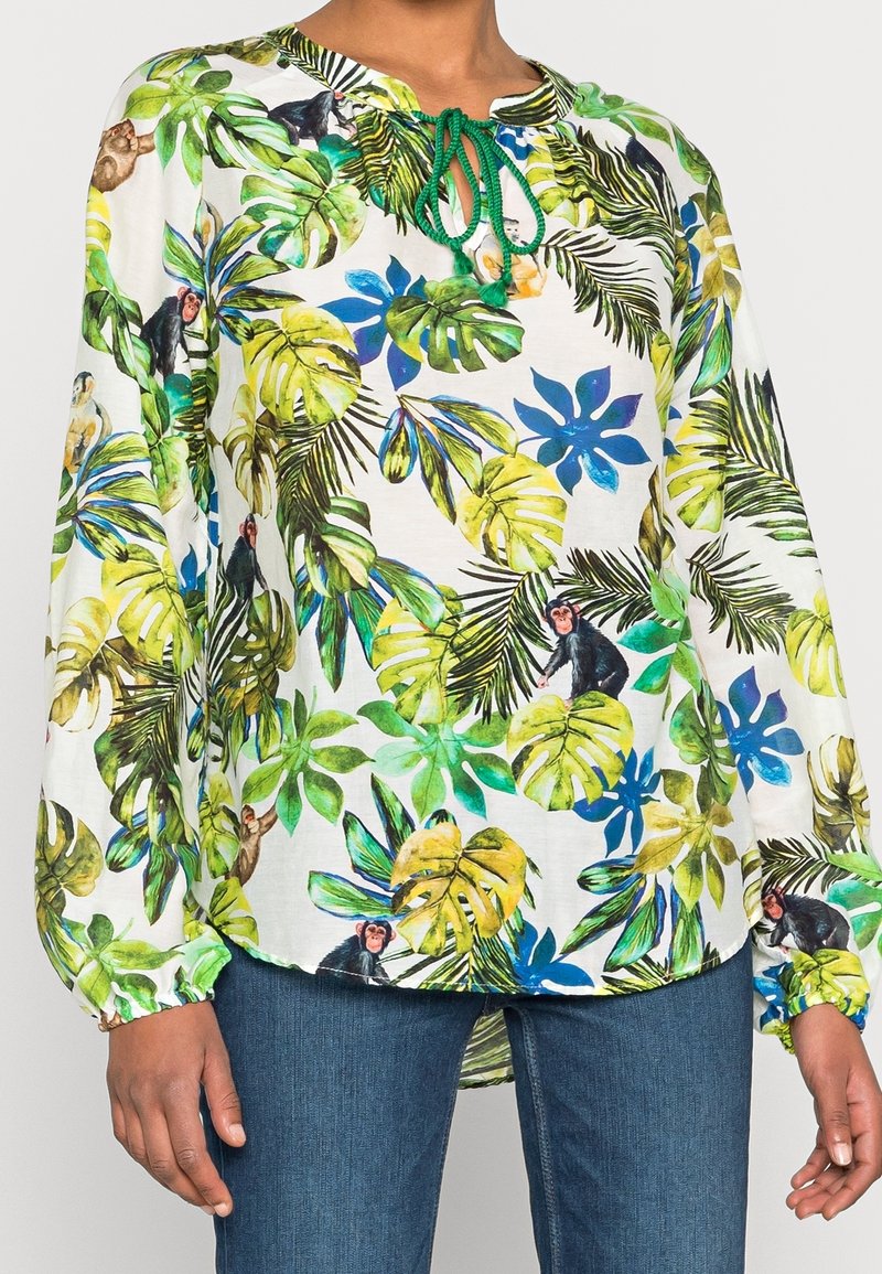 Blouse blanche à manches longues avec un ruban vert au cou, ornée de feuilles tropicales colorées et de petites illustrations de singes, portée avec un jean bleu.