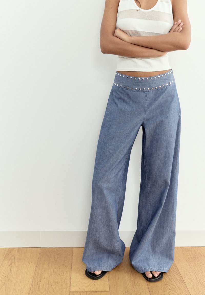 Personne debout, bras croisés, portant un pantalon en jean bleu à taille haute et jambes larges avec des clous métalliques et un haut blanc sans manches.