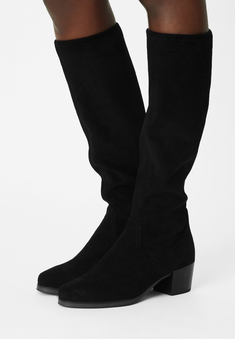 Bottes hautes noires en daim avec un petit talon carré portées sur des jambes nues sur un fond clair uni.