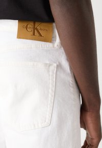 Bijele traperice od denima s kožnom oznakom Calvin Klein Jeans na leđima. Značajke uključuju ravni džep i vidljivo šivanje.