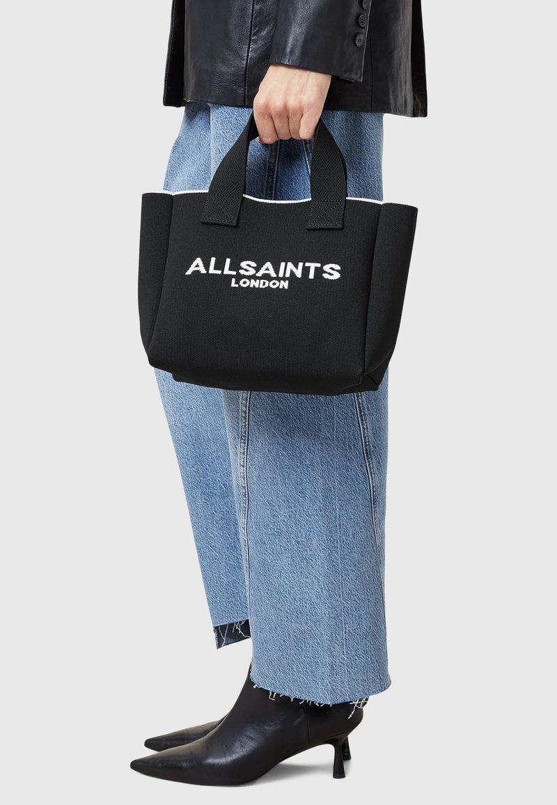 Borsa tote nera con il testo bianco "ALLSAINTS LONDON". Presenta due manici e un design robusto. Indossata con jeans blu a gamba larga e tacchi neri.