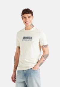 T-shirt di cotone color crema con una grafica blu e la scritta "GUESS EST. 1981" sul davanti. Il design include un bordo testurizzato e maniche corte.