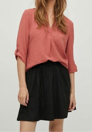 Femme portant un chemisier saumon-rose texturé avec manches retroussées et une jupe noire à cordon, sur un fond uni.