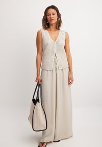 Haut sans manches en maille côtelée beige avec un décolleté en V et une fermeture à l'avant. Associé à un pantalon large crème et à un sac cabas beige.