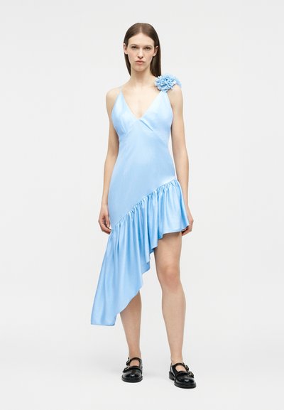 Γυναίκα που φοράει ένα ανοιχτό μπλε ασύμμετρο φόρεμα με λεπτομέρεια λουλουδιού στον ώμο και μαύρα παπούτσια με αγκράφα, στέκεται μπροστά σε ένα απλό φόντο.