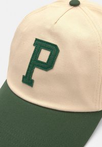 Căciulă bej cu margine verde, având un „P” verde brodat pe partea din față. Fabricată dintr-un material texturat cu o coroana rotundă.