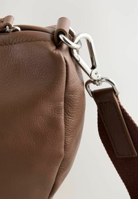 Braune Lederhandtasche mit sichtbaren Nähten, runder Form, silberner Hardware und einem Stoffgriff mit strukturierter Oberfläche.