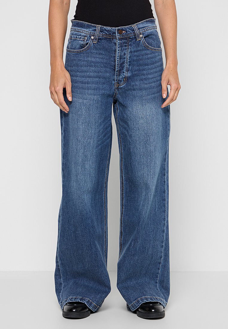 Person mit weit geschnittenen blauen Jeans, die sichtbare Fronttaschen haben, und schwarzen Schuhen, steht vor einem einfarbigen Hintergrund.