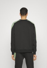 Mężczyzna odwrócony plecami, ubrany w czarną bluzę i dopasowane spodnie z jasnoszarymi paskami na rękawach i bokach spodni, stojący na białym tle.